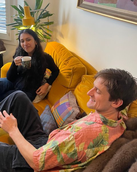 Tommy Antonio and Mallika Bhagwat relaxing on a yellow sofa with a mug of tea